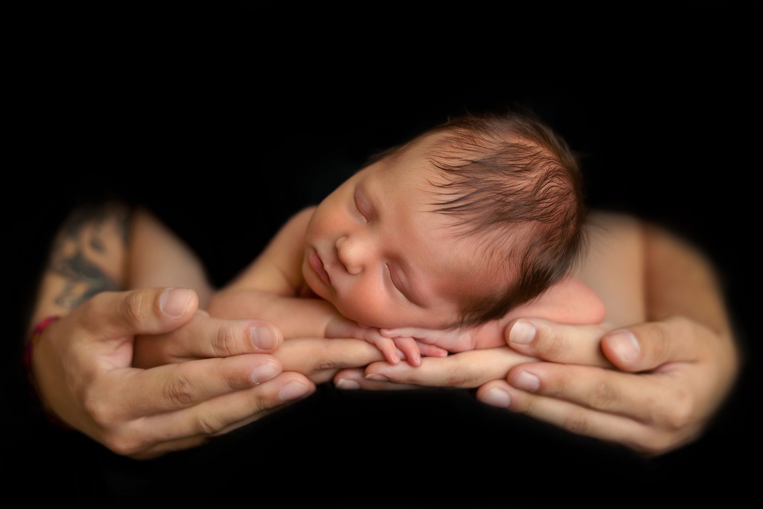 Artistic portrait of sleeping newborn cradled in gentle hands against black background.