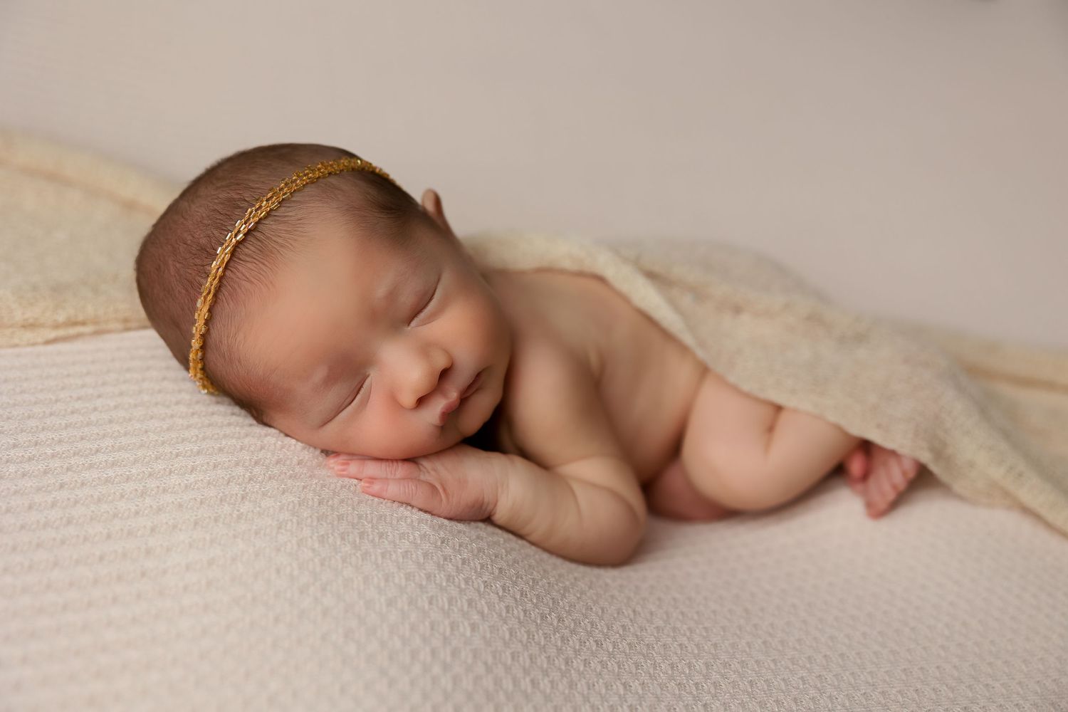 Peaceful newborn sleeping on textured cream fabric with delicate gold headband.