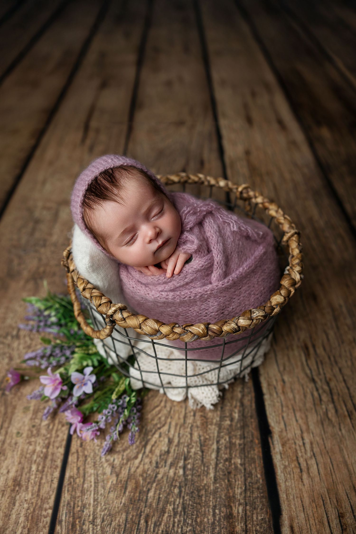 Sleeping newborn wrapped in purple fabric nestled in woven basket with flowers on wooden floor.