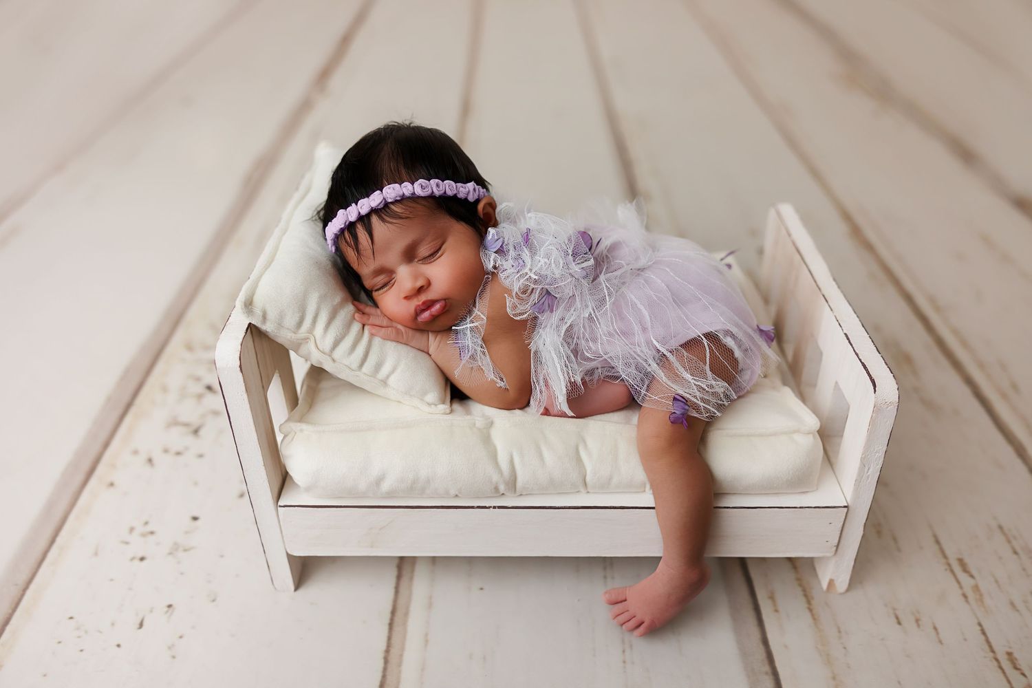 Peaceful sleeping newborn wearing a delicate headband rests on white bedding in a small wooden bed.