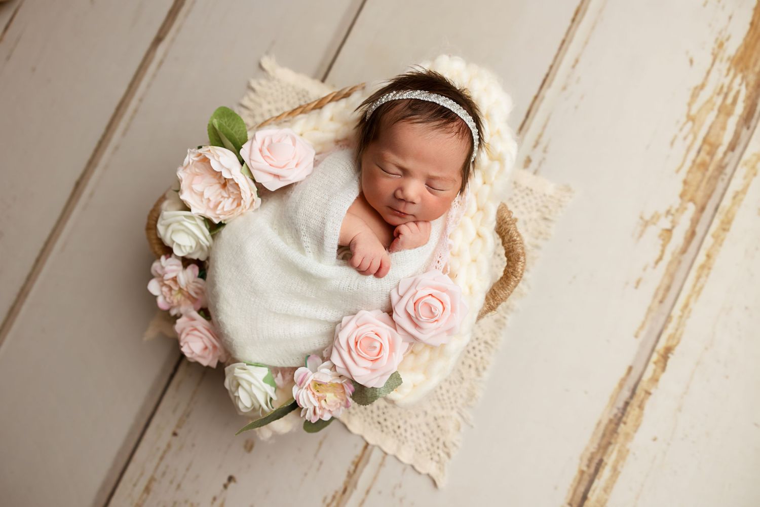 Sleeping newborn wrapped in white with pink roses on rustic wooden backdrop.