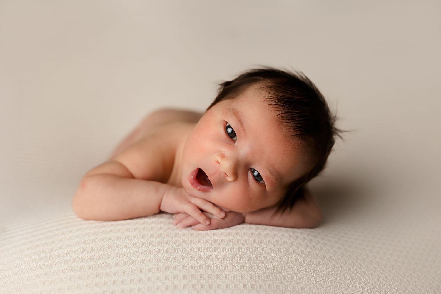 Awake newborn lying on cream textured surface with hand near mouth.