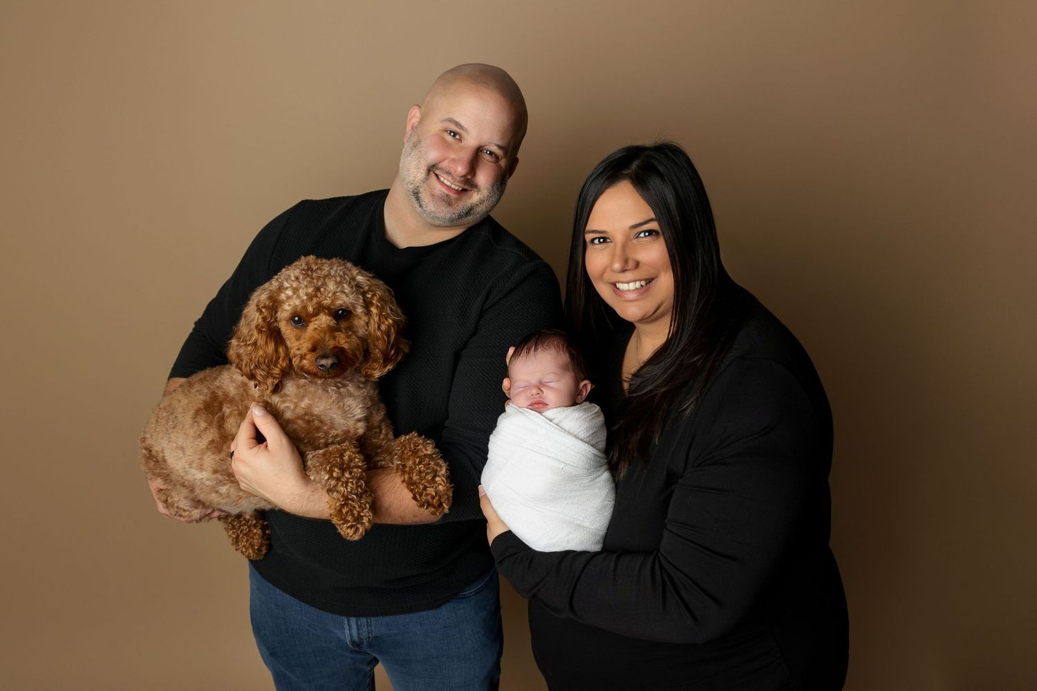 Family portrait with goldendoodle dog and swaddled baby against brown backdrop.