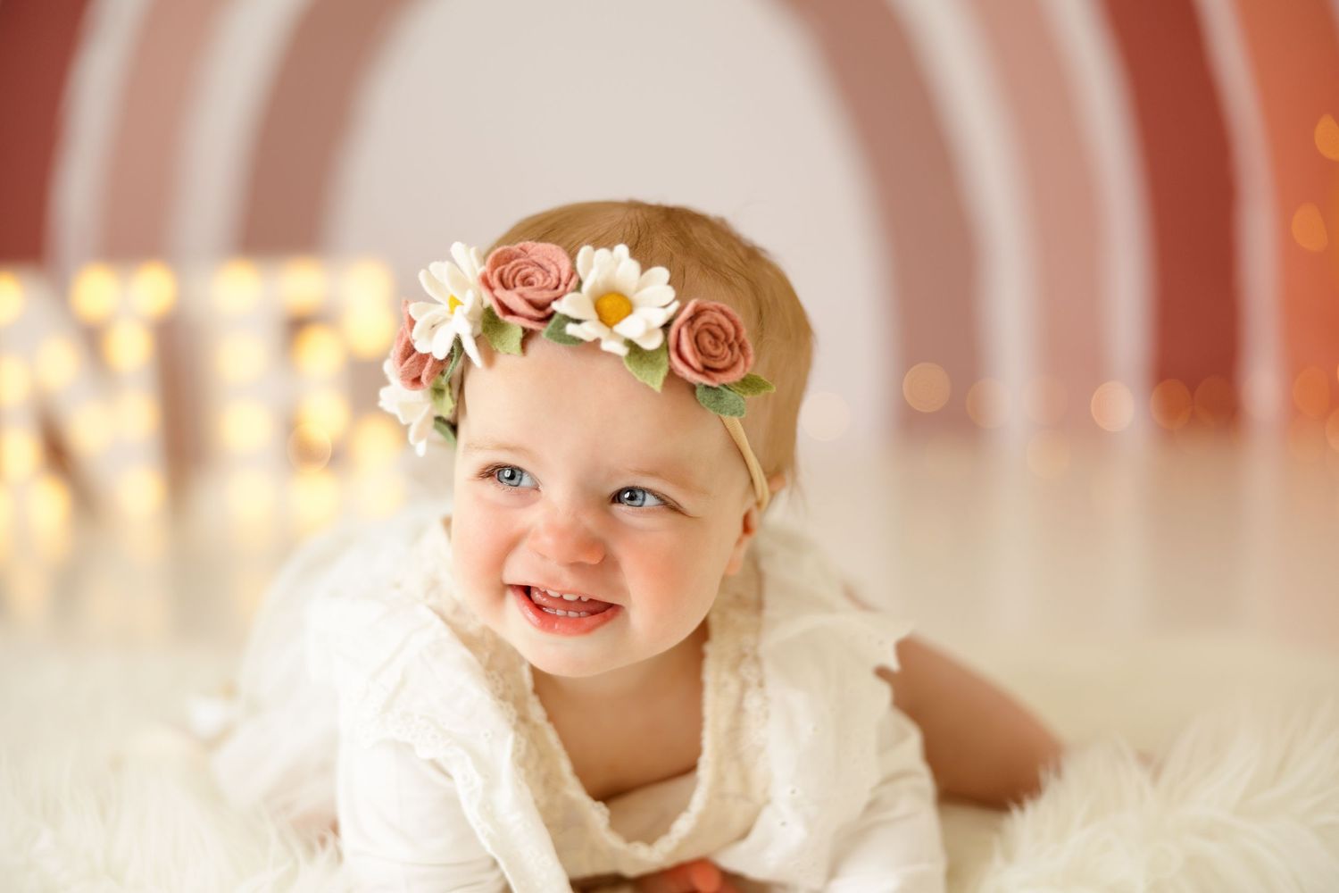 Happy baby wearing floral crown lying on white fur against pink circular background.