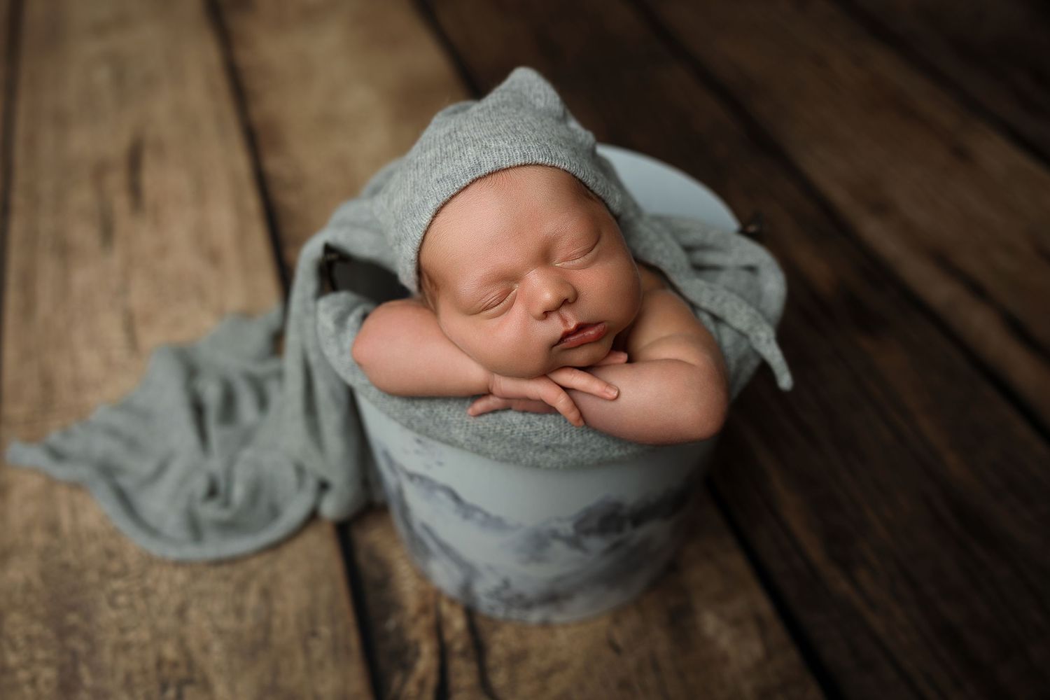 Newborn baby sleeping peacefully in a teal wrap and bonnet inside a bucket on rustic wood floors.