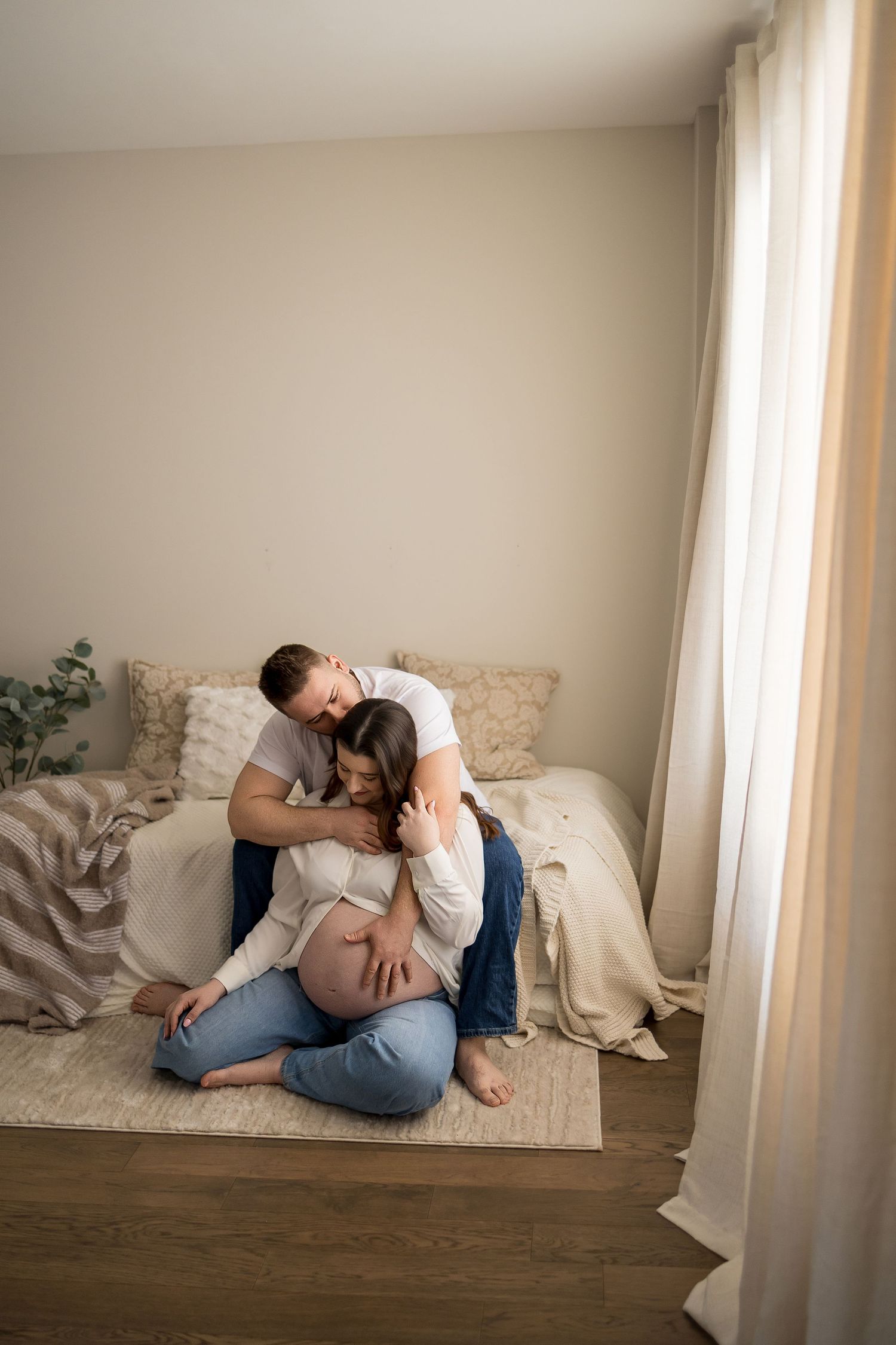A couple shares an intimate embrace while sitting on the floor of a bright room with white curtains and hardwood floors.