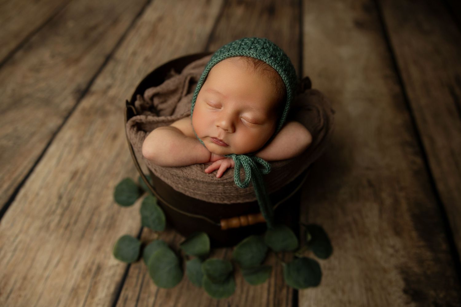 Sleeping newborn in green bonnet rests in wooden bowl with eucalyptus decor.
