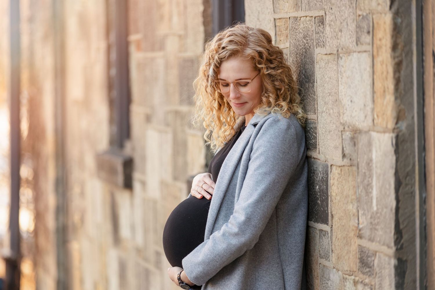 Maternity photo of woman in grey coat leaning against stone wall.
