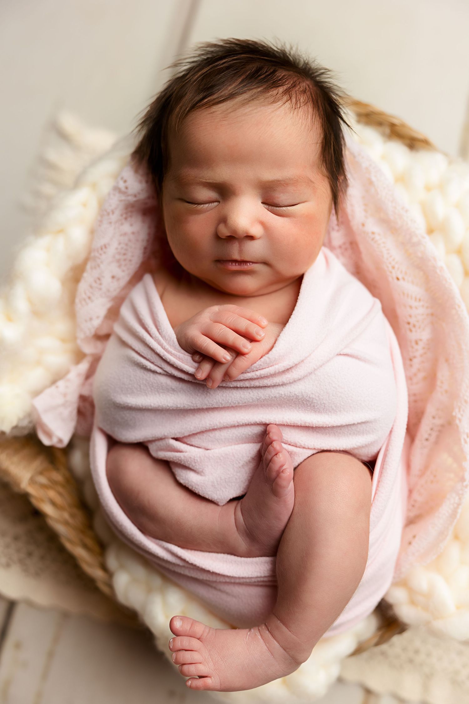 Sleeping newborn wrapped in pink swaddle on cream textured backdrop.