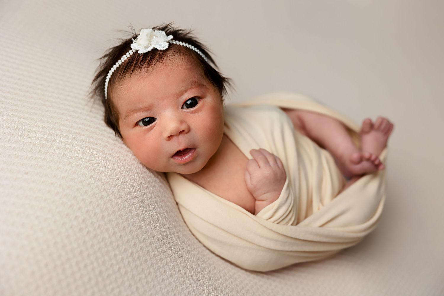 Peaceful newborn baby swaddled in cream wrap on white textured surface with flower hair accessory.