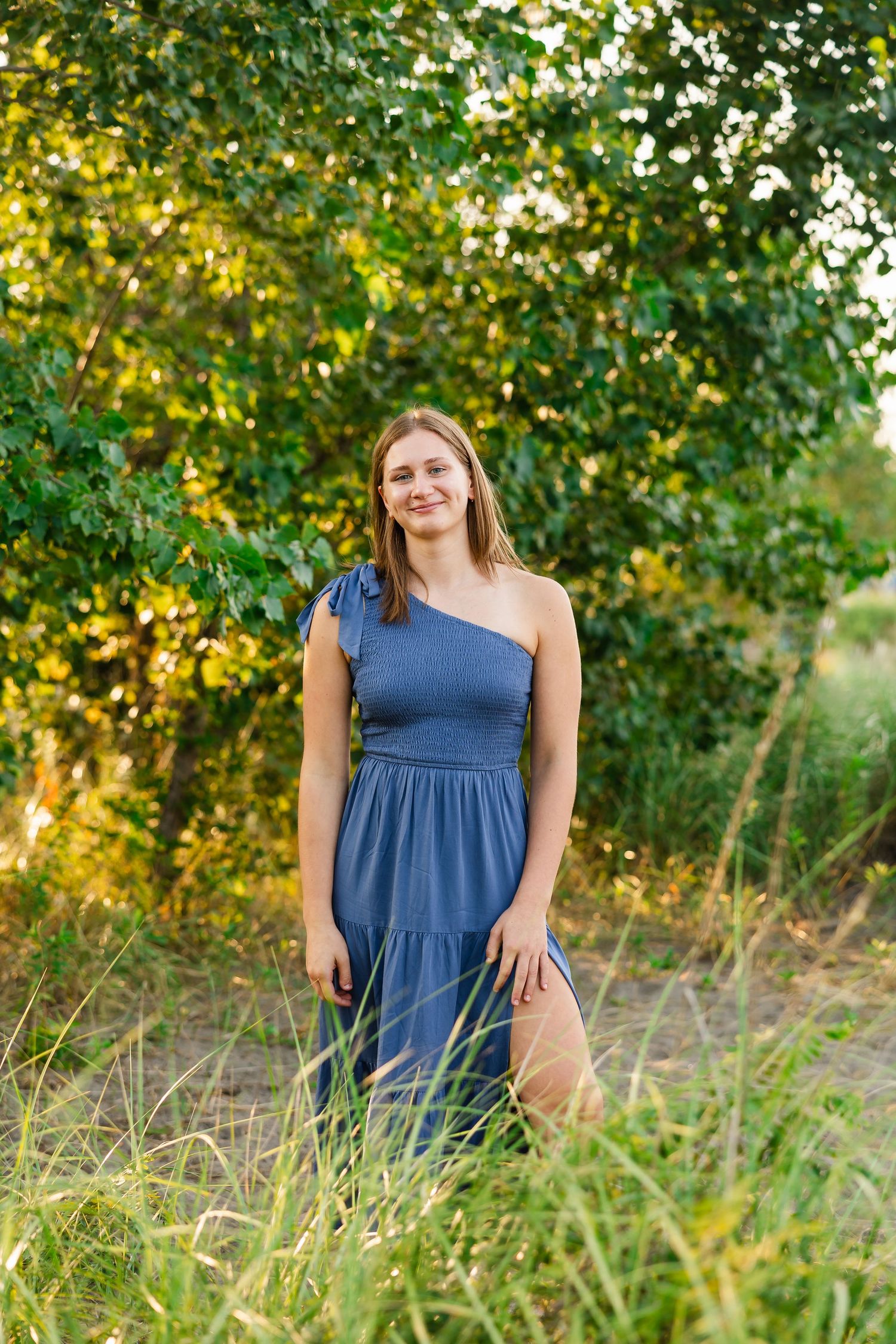 Woman in blue one-shoulder dress stands in tall grass with sunlit trees in background during golden hour.