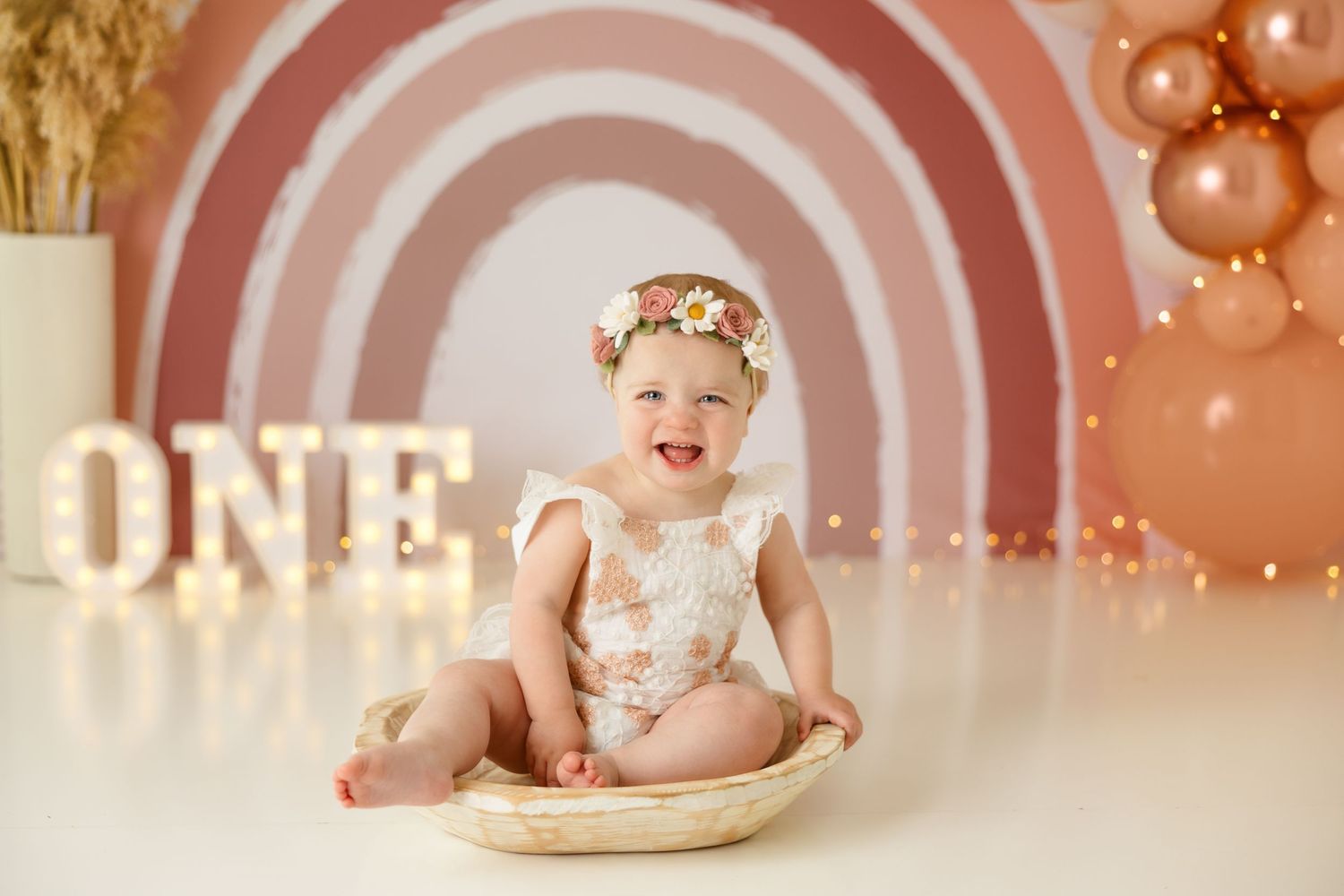 Baby in floral romper sitting in wooden bowl against pink backdrop with 'ONE' lettering.