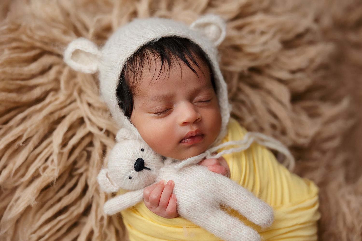 Sleeping newborn in yellow wrap and white bear bonnet on fur backdrop.