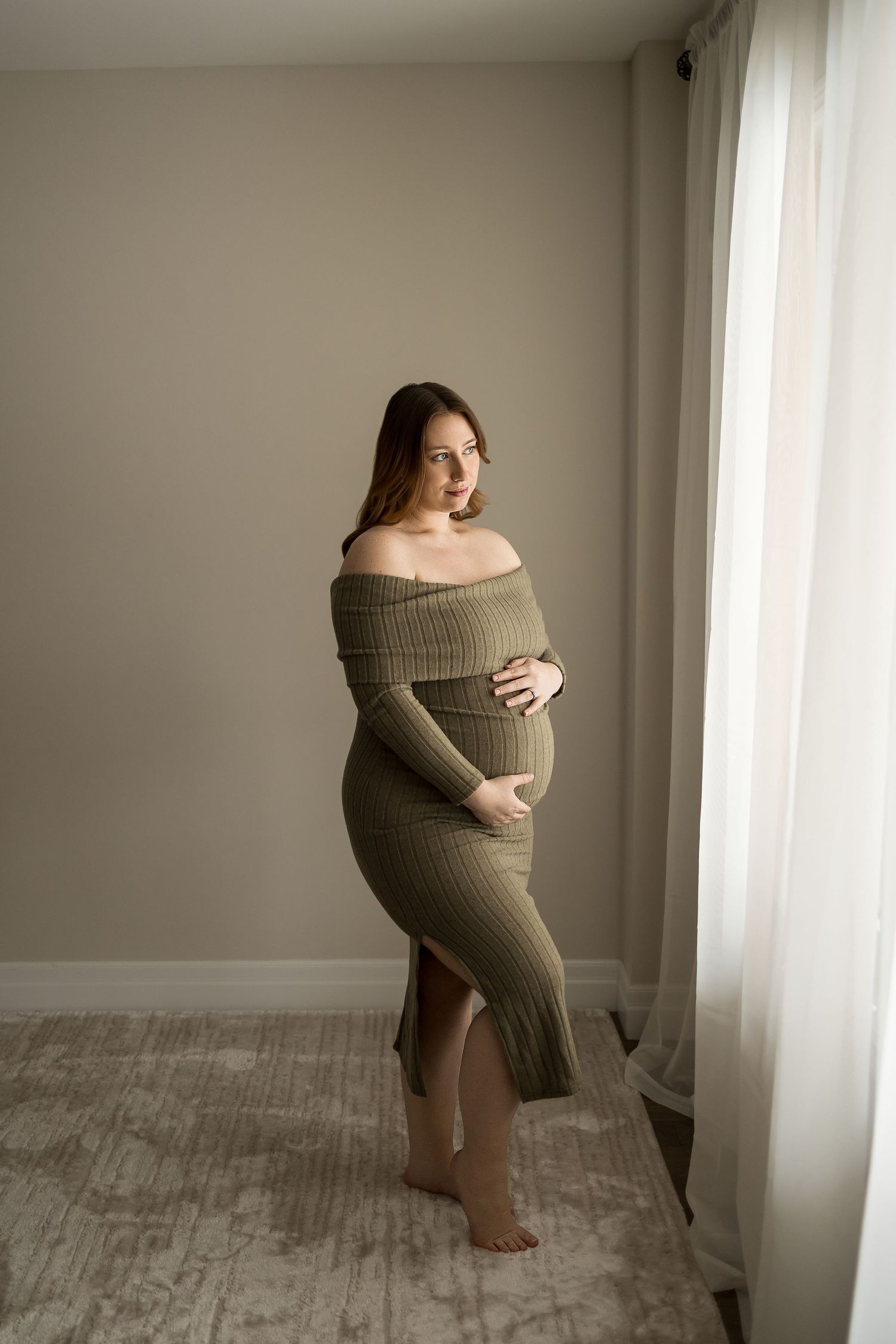 Artistic maternity photos showing an expectant mother in a beige sweater dress by a window in natural light.
