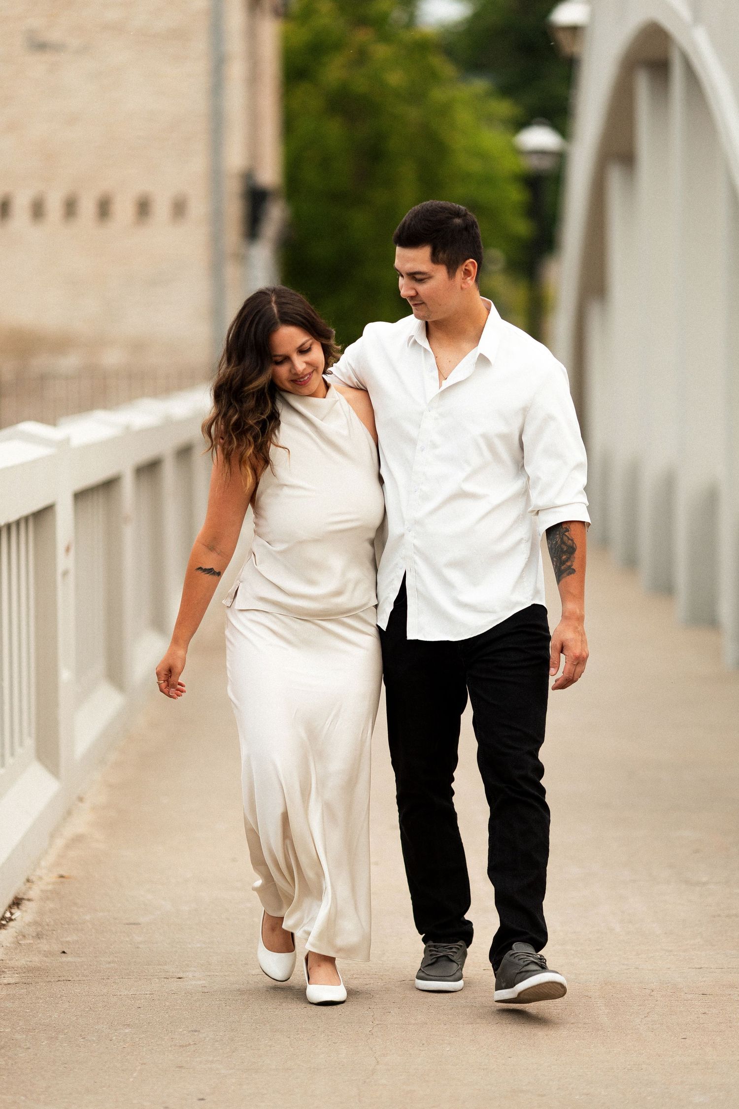 Couple walking arm in arm on a bridge, woman in white dress and man in white shirt with black pants.