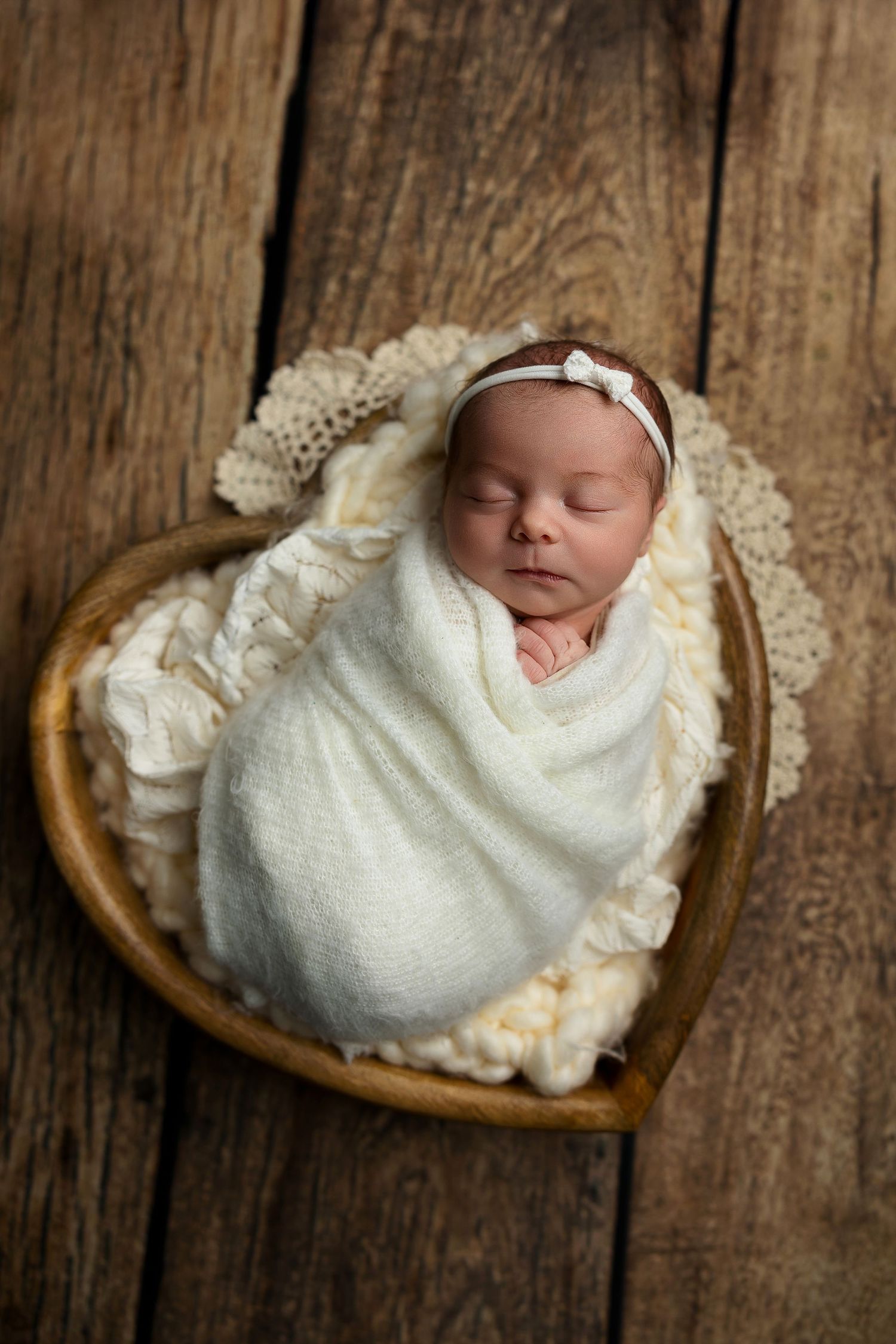 Sleeping newborn baby wrapped in white blanket lies in heart-shaped bowl on rustic wooden surface.