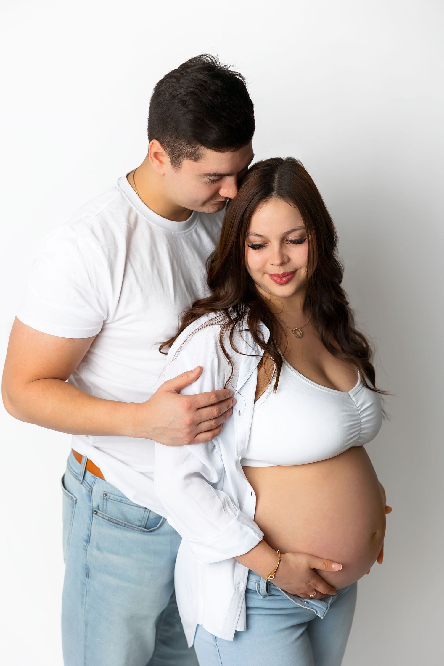 Intimate maternity photoshoot featuring couple in casual white attire against white backdrop.