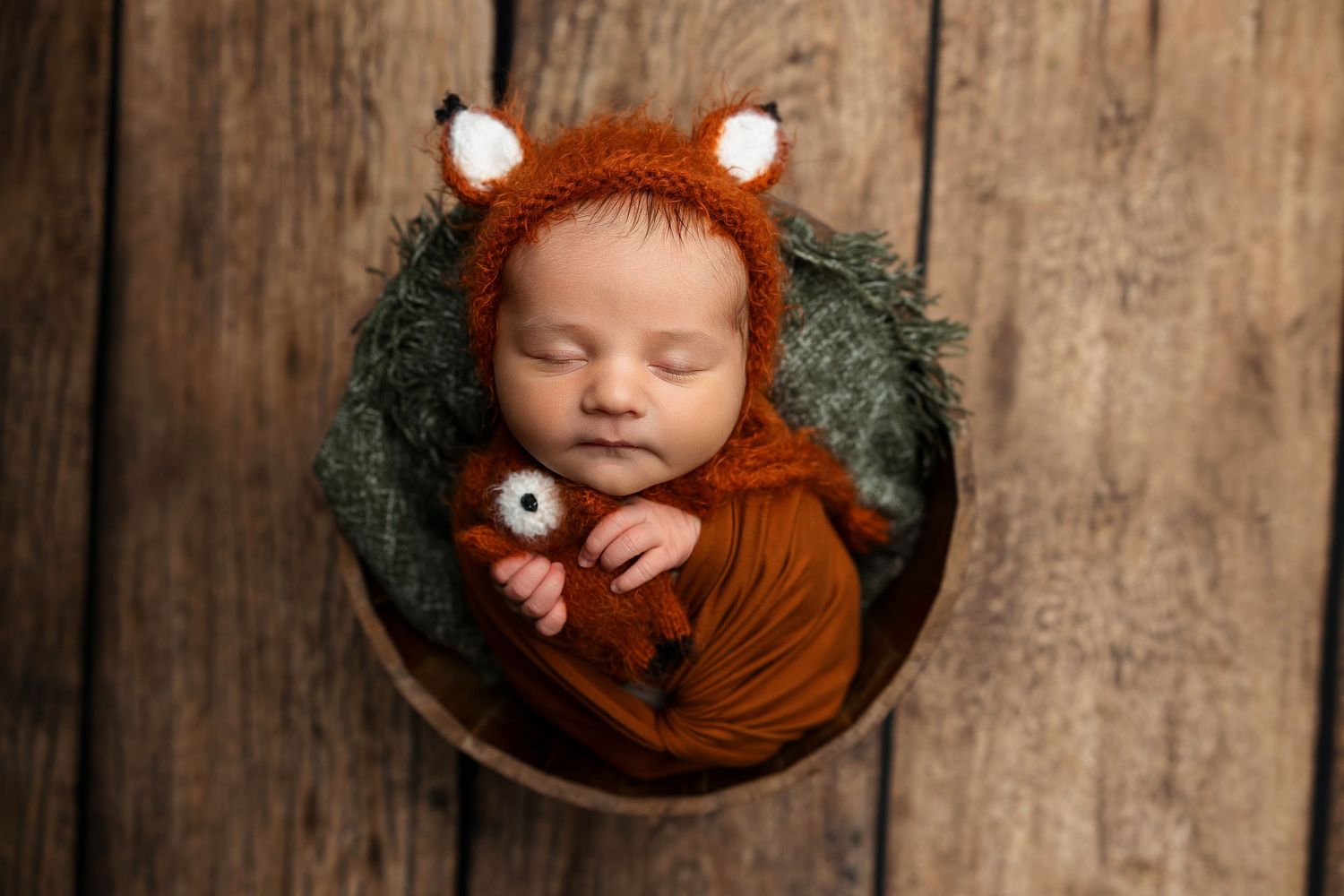 Newborn wearing fox-themed bonnet sleeping peacefully in round basket.