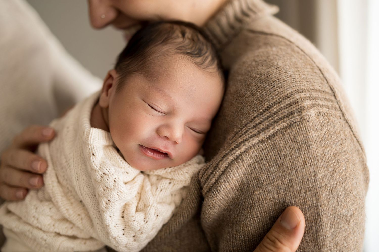 Newborn baby swaddled in cream wrap sleeping peacefully in parent's arms wearing a brown sweater.