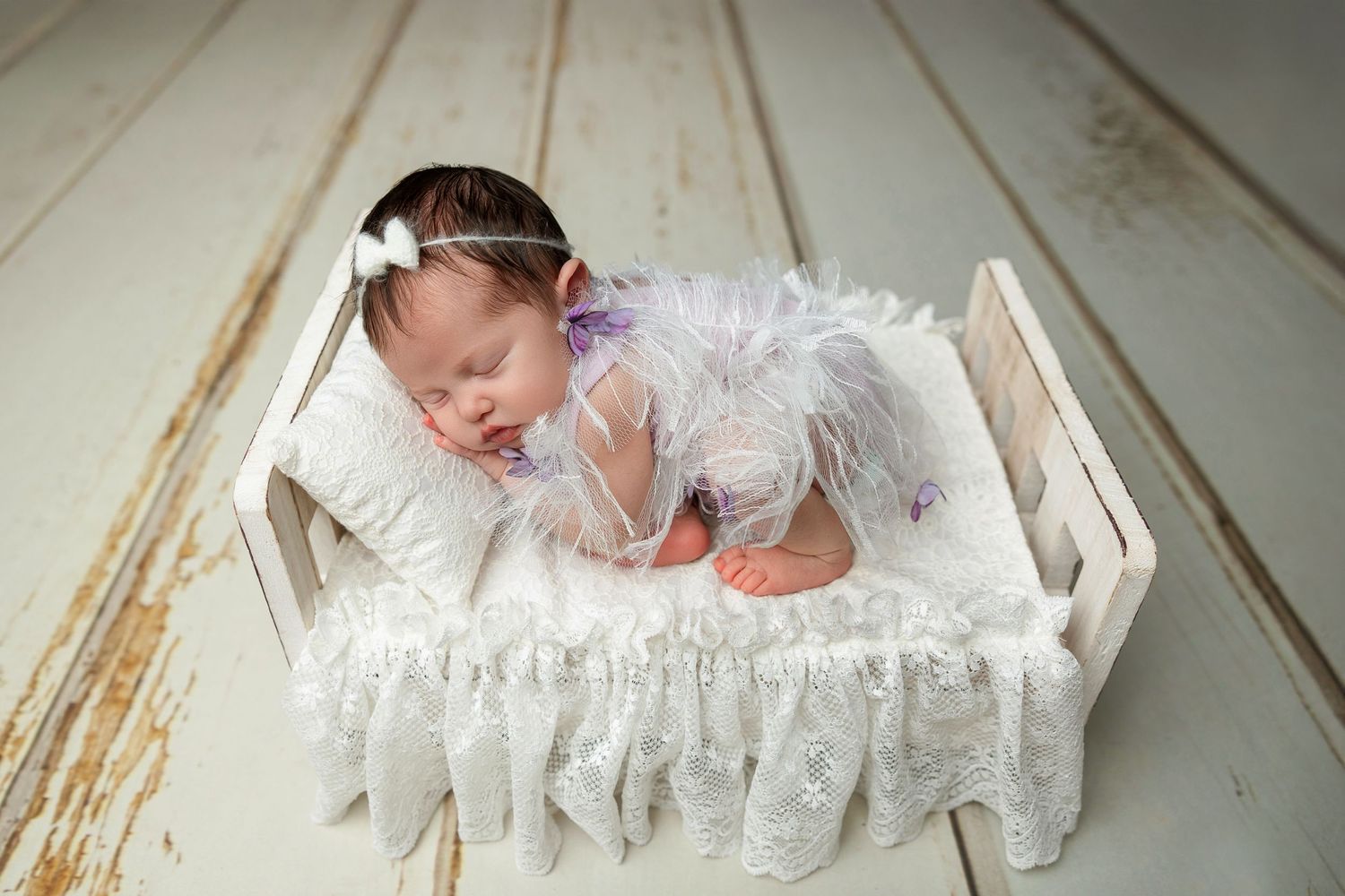 Sleeping baby in white lace outfit resting in rustic wooden bed with delicate fabric details.