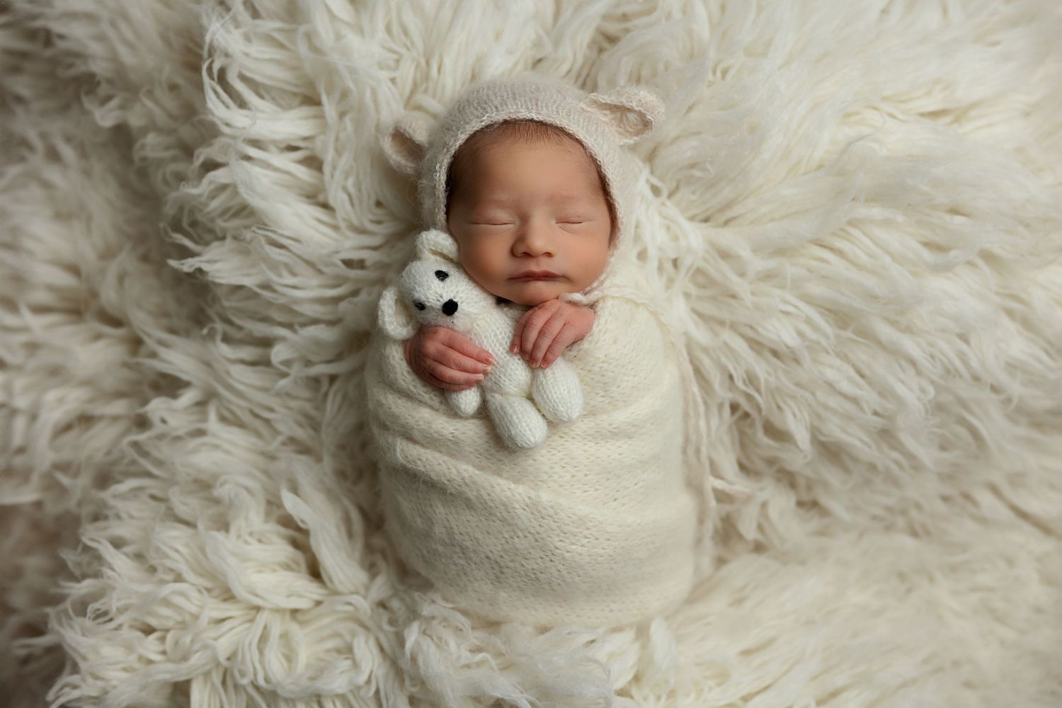 Sleeping newborn with tiny plush toy wrapped in white on furry backdrop.