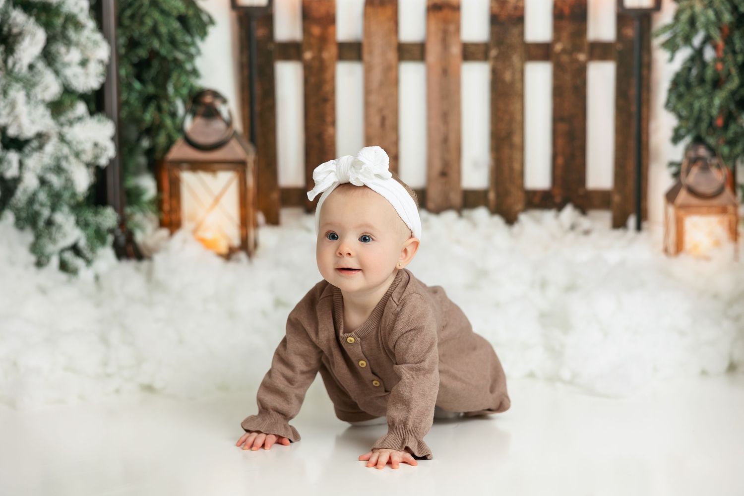 Baby in brown outfit crawling on snowy white floor with holiday decorations.