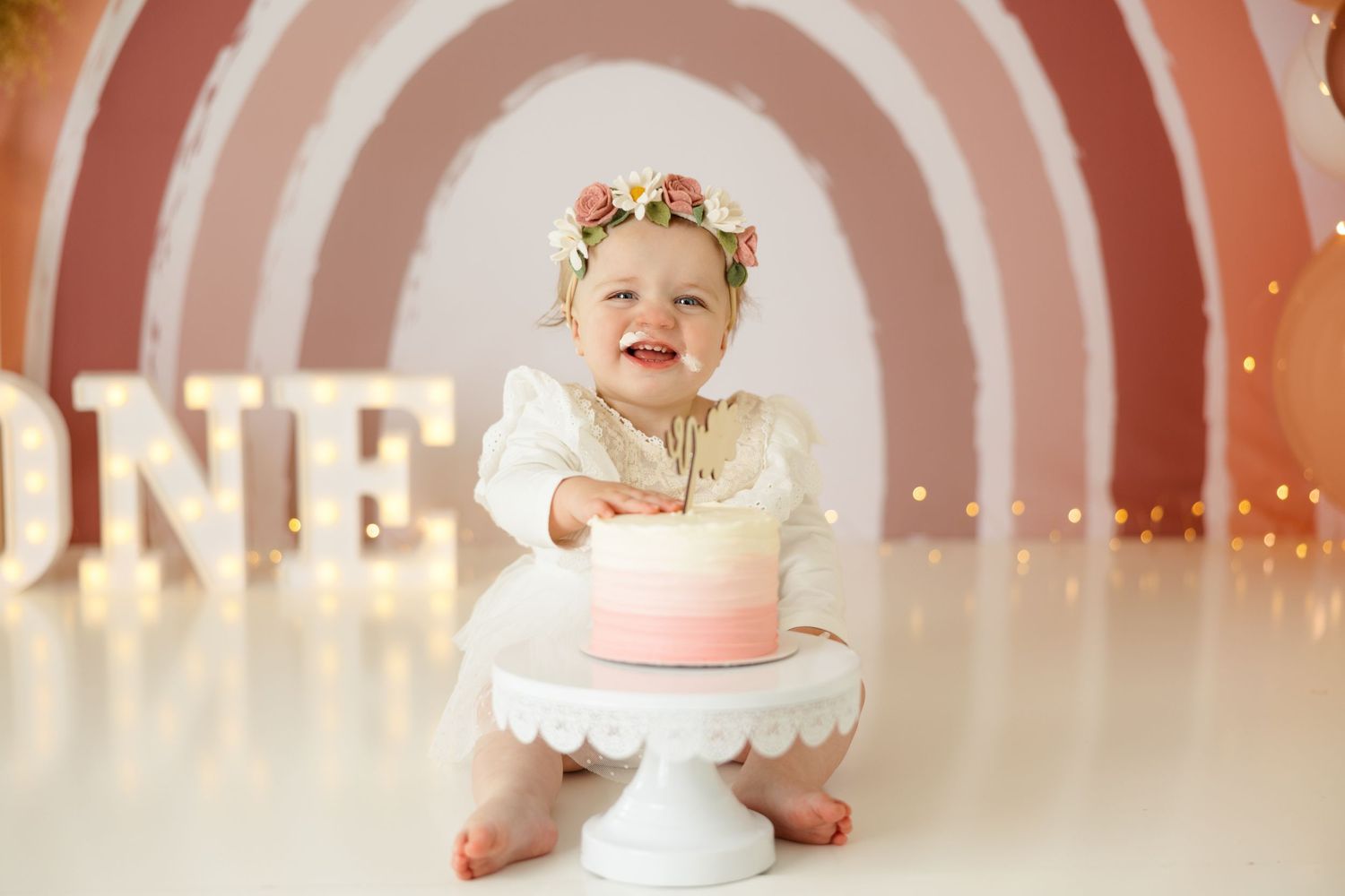 Cake smash photo session with circular rainbow backdrop and rose gold balloon decorations.