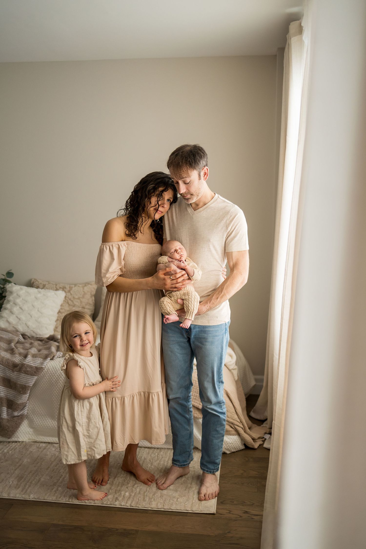 A family stands together by a bright window in their home wearing neutral tones and casual clothing.
