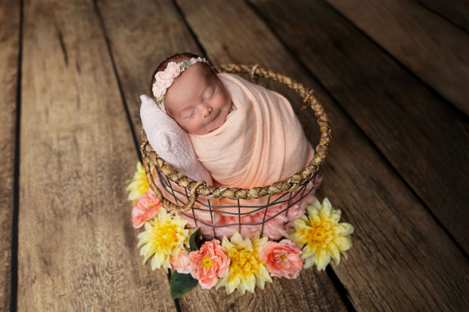 Sleeping newborn nestled in floral basket arrangement on rustic wooden floor.