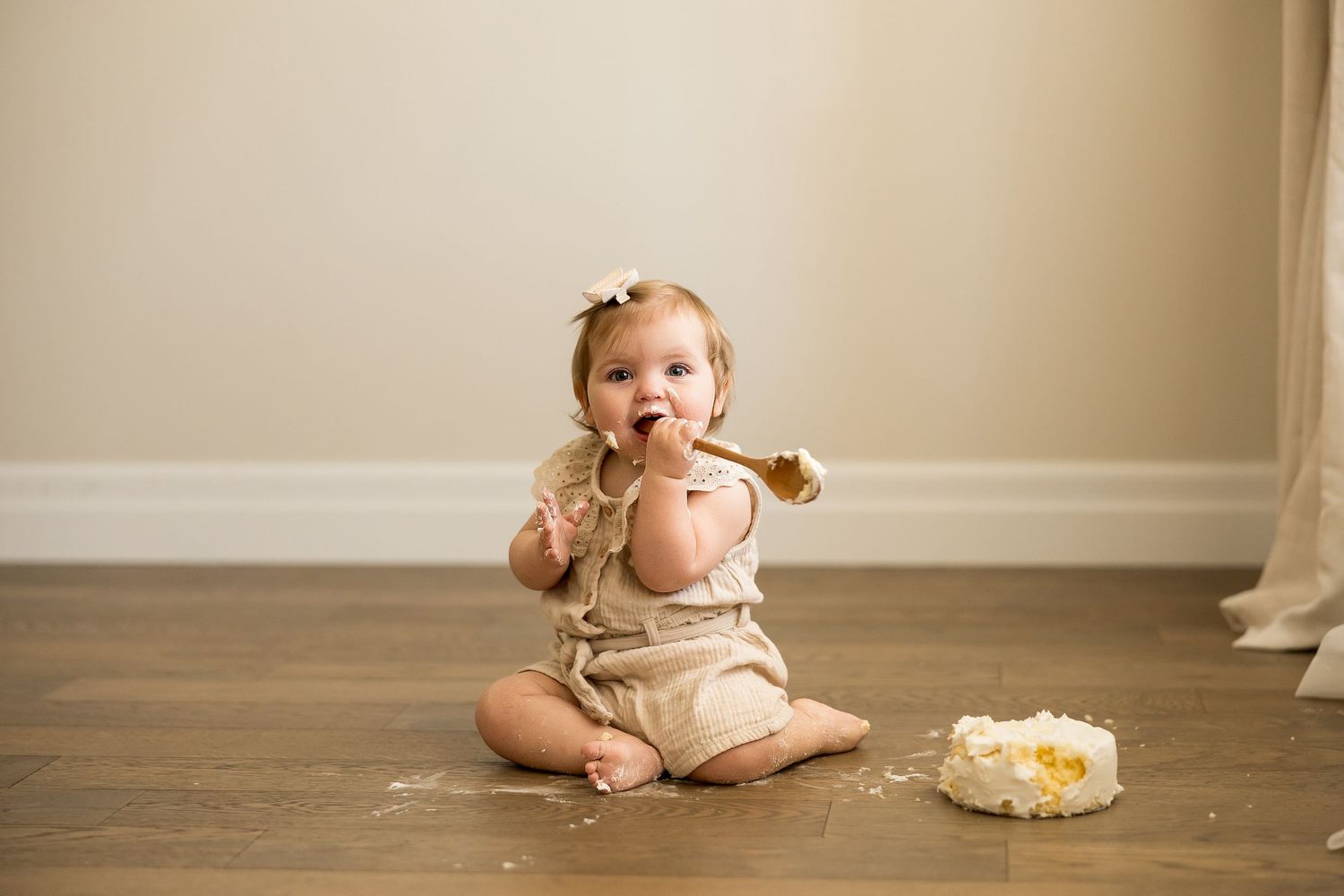 Baby girl in beige romper sitting on hardwood floor eating cake with spoon during first birthday smash cake session.