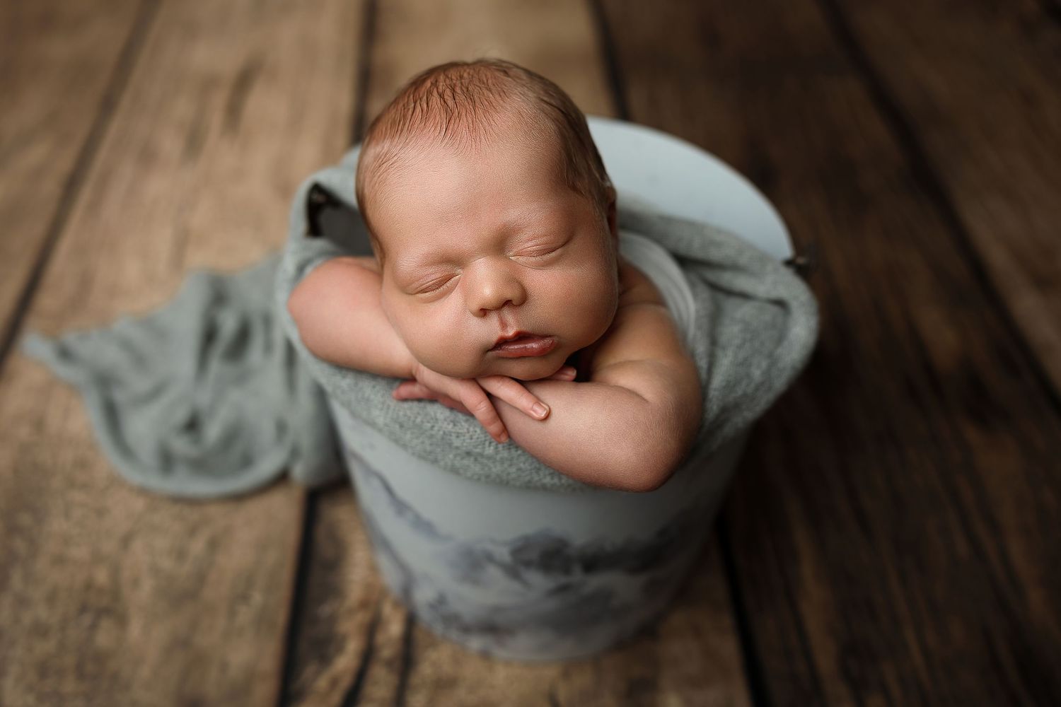 Sleeping newborn baby wrapped in soft blue fabric inside a bucket on rustic wood floor.