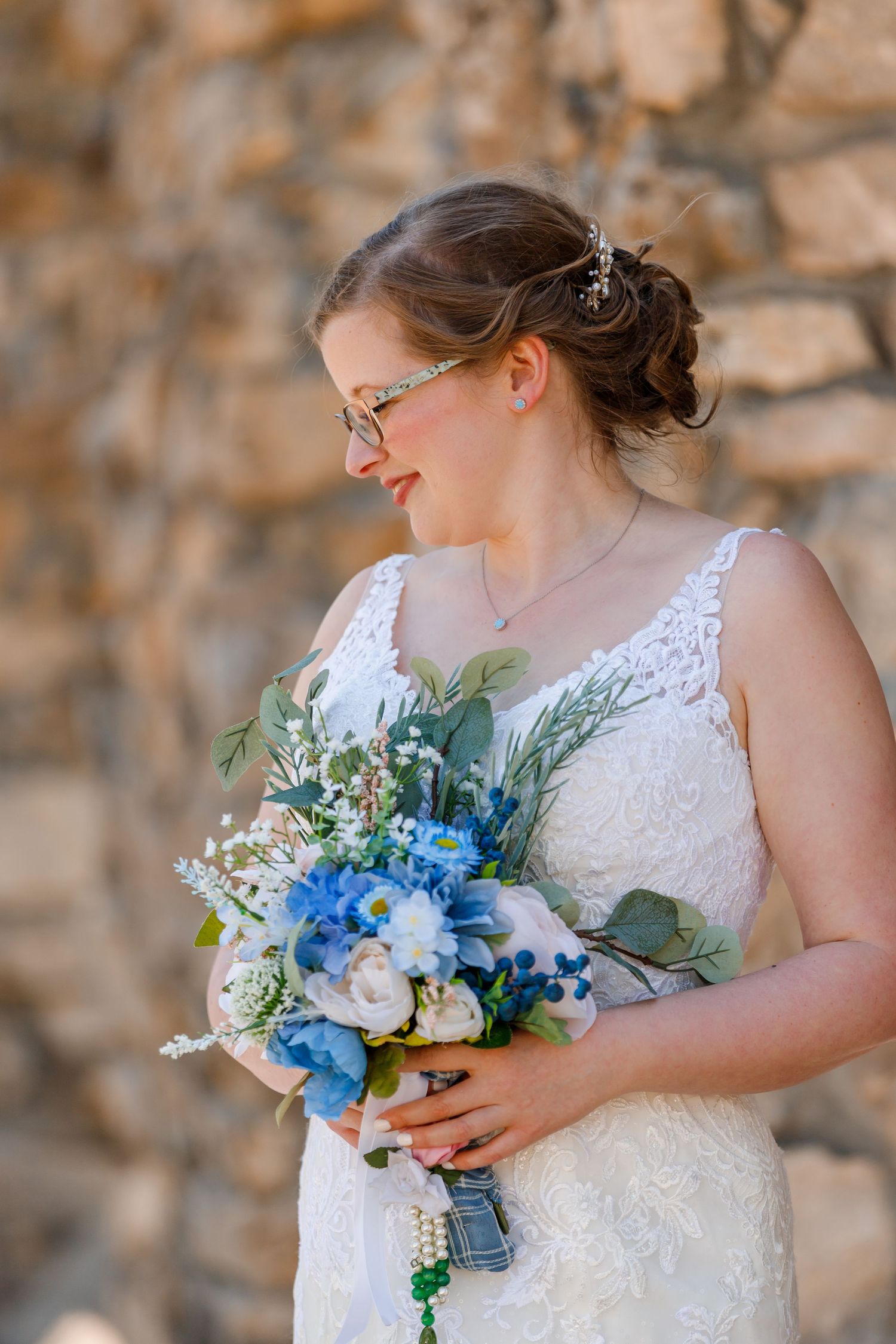 Bride holding vibrant blue and white bouquet wearing lace dress and glasses against stone wall.