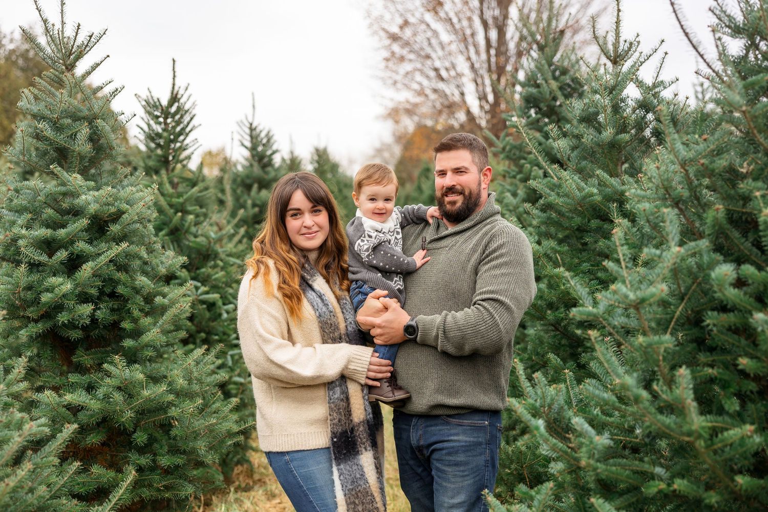 Family of three posing together at a Christmas tree farm surrounded by evergreen trees.
