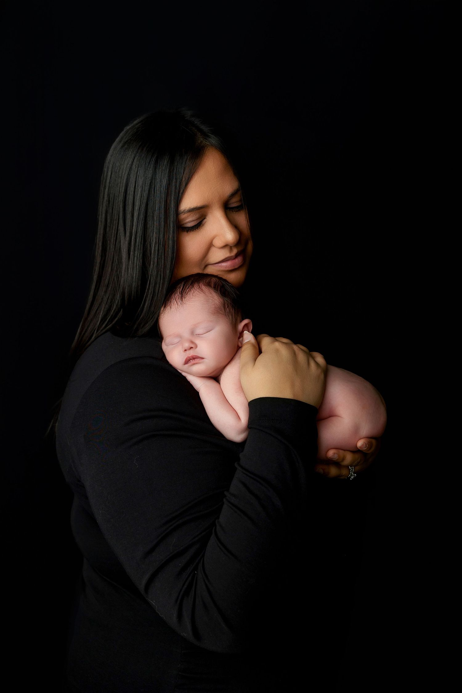A tender moment captured in a dark studio as a woman in black cradles a sleeping newborn against her chest.