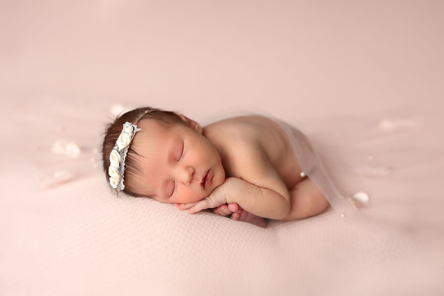 Peaceful newborn wearing lace headband sleeping on soft pink background.