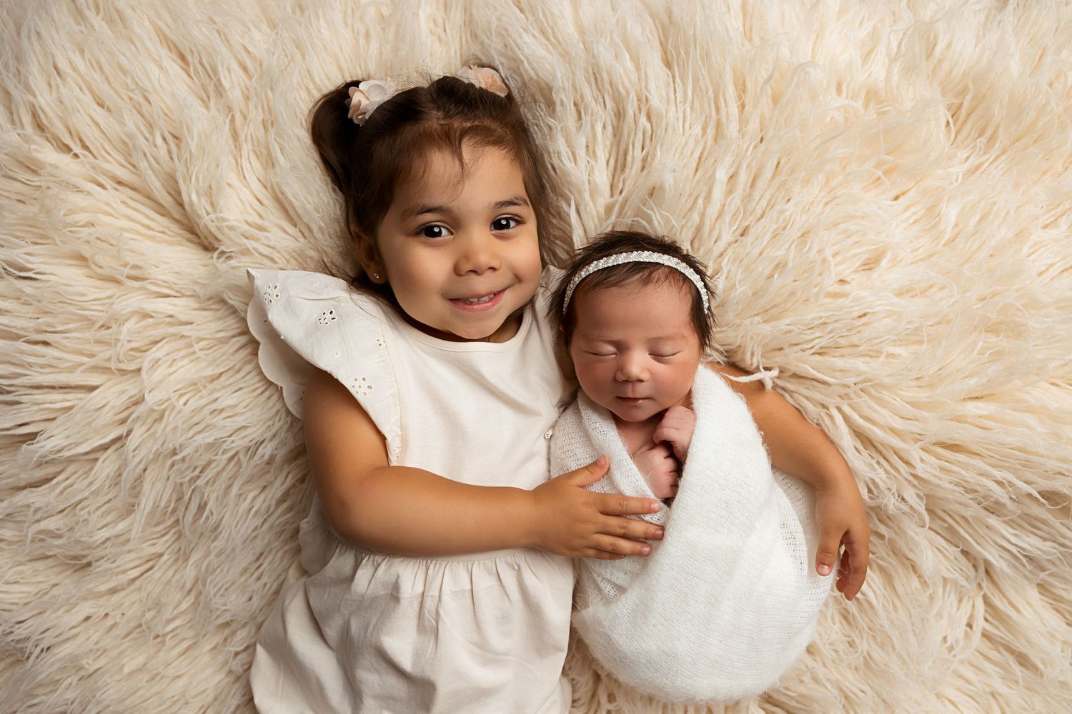 Sisters in white dresses snuggling on plush cream fur backdrop.