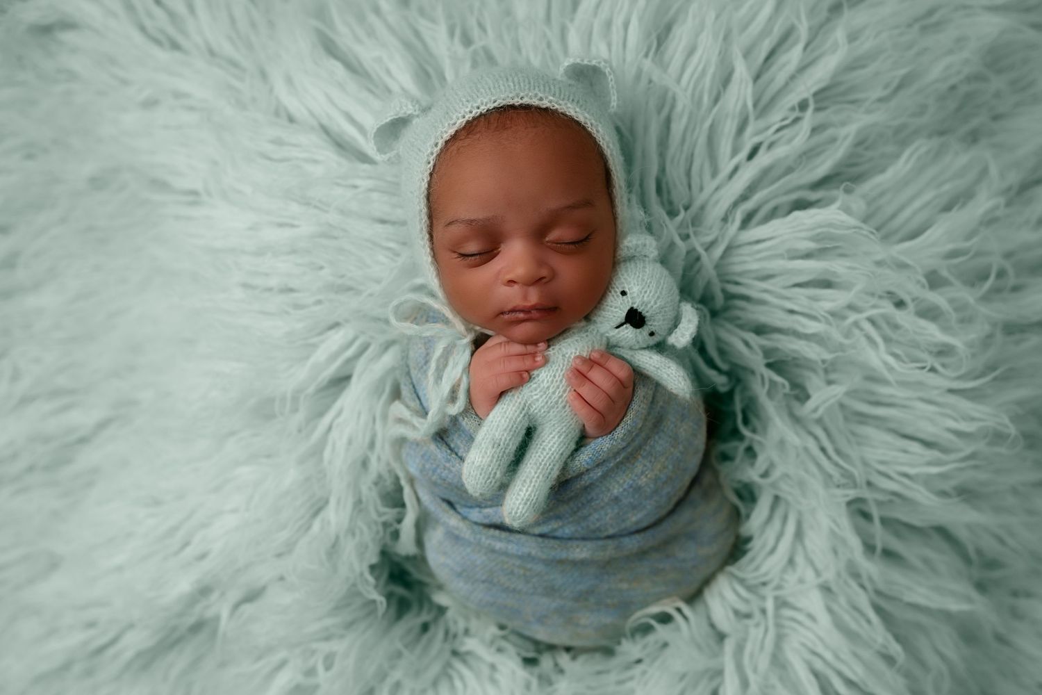 Sleeping newborn wrapped in soft blue swaddle on plush mint-colored fur blanket.