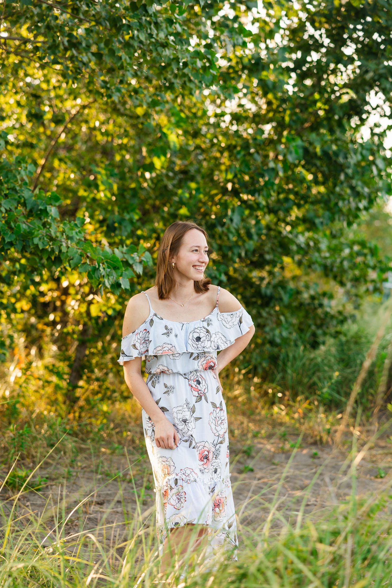A soft floral off-shoulder dress is showcased in an outdoor setting with golden sunlight filtering through green trees.