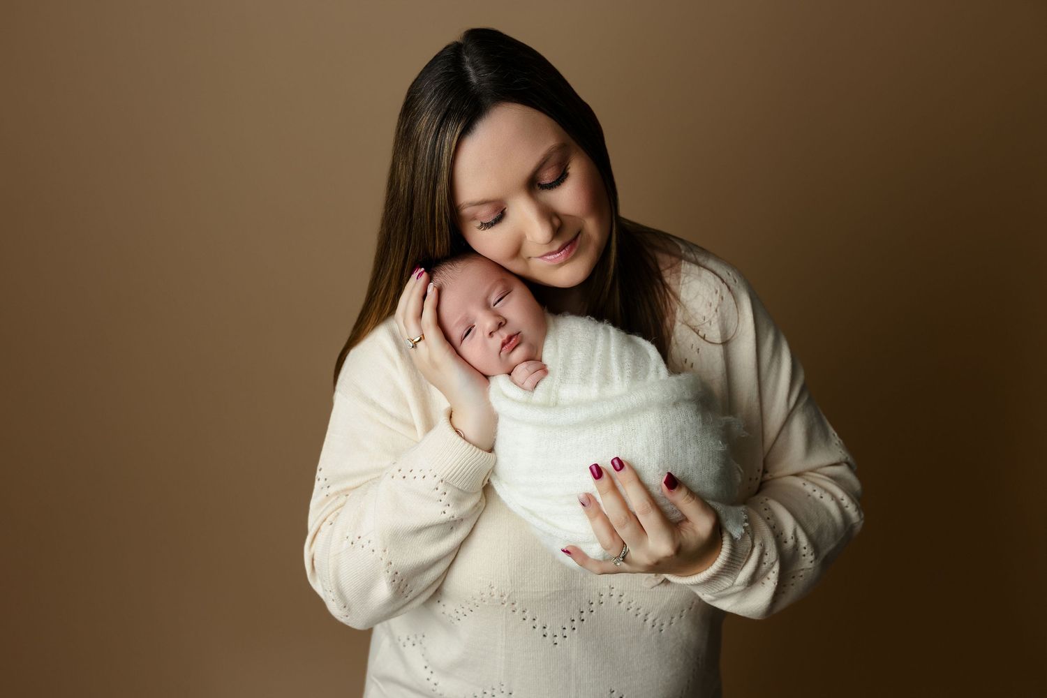 A tender moment between parent and newborn baby against a brown backdrop, both wearing cozy white sweaters.