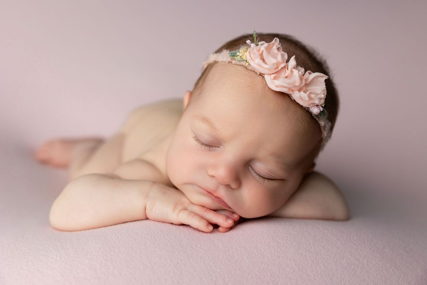 Peaceful newborn sleeping on soft pink fabric with delicate lace headband in professional studio portrait.