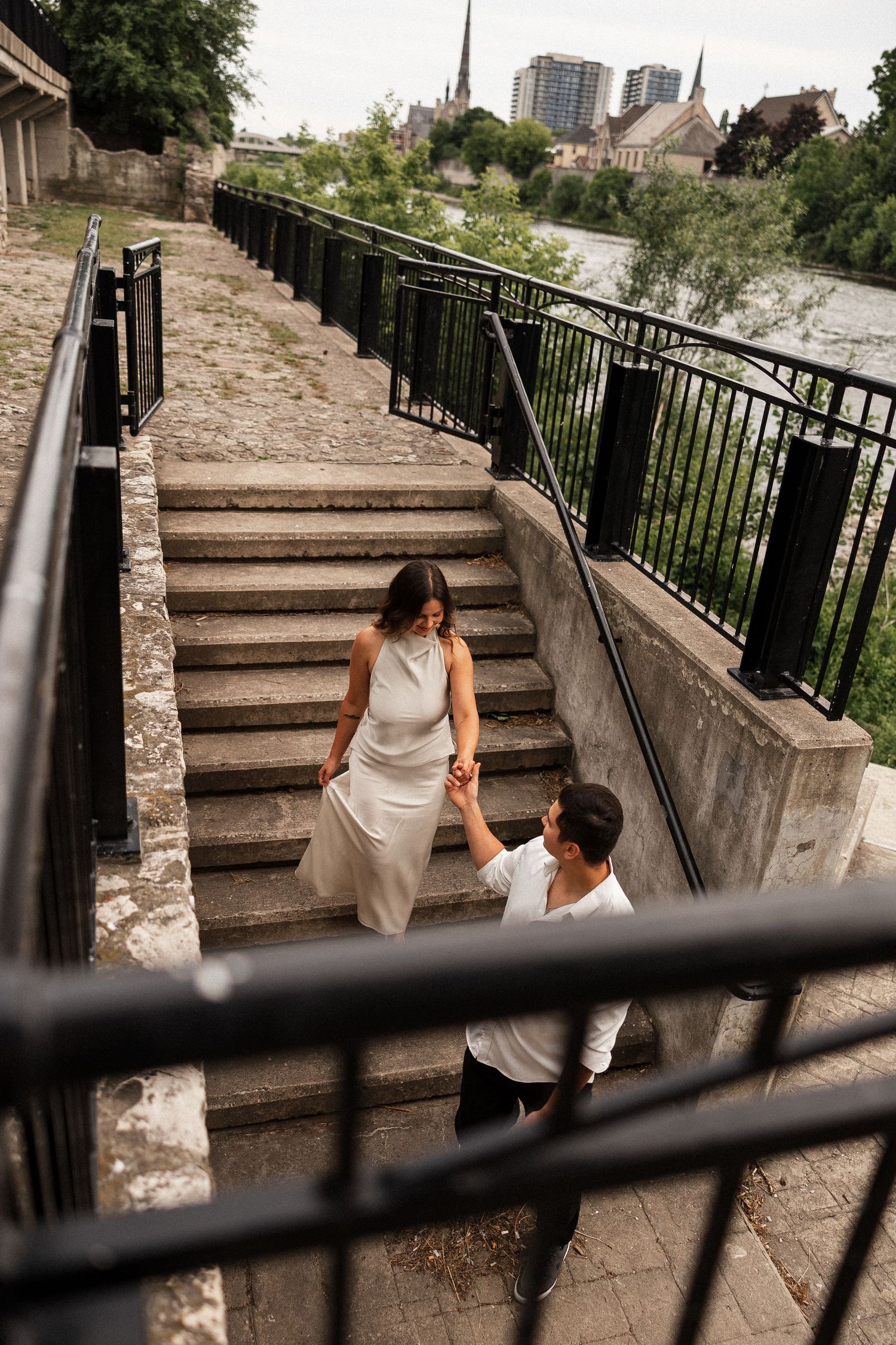 Couple holding hands on stone steps by a river, woman in white dress leads man upward in a romantic urban outdoor setting.