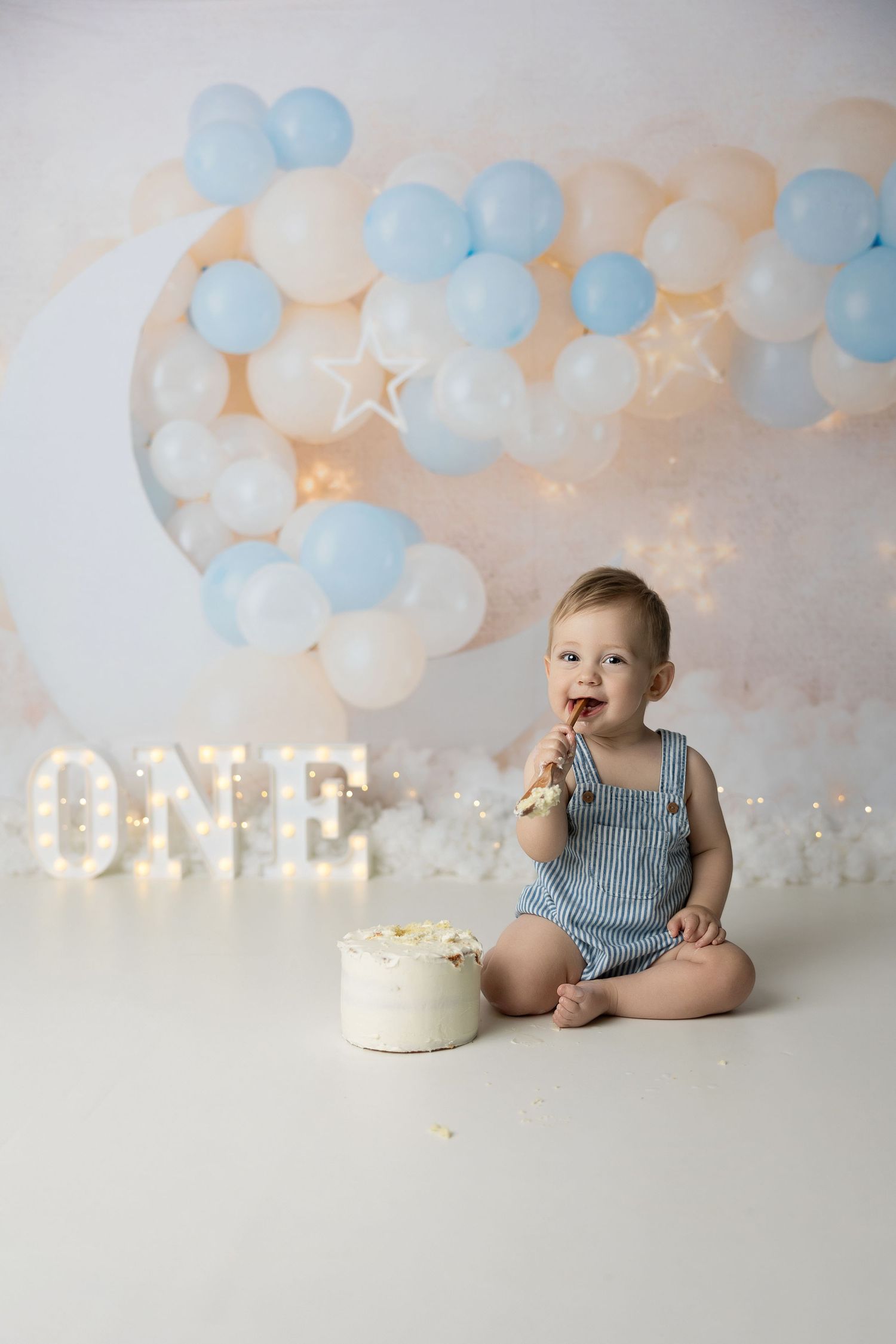 A festive first birthday setup with illuminated ONE sign, blue balloons, and white cake on light backdrop.
