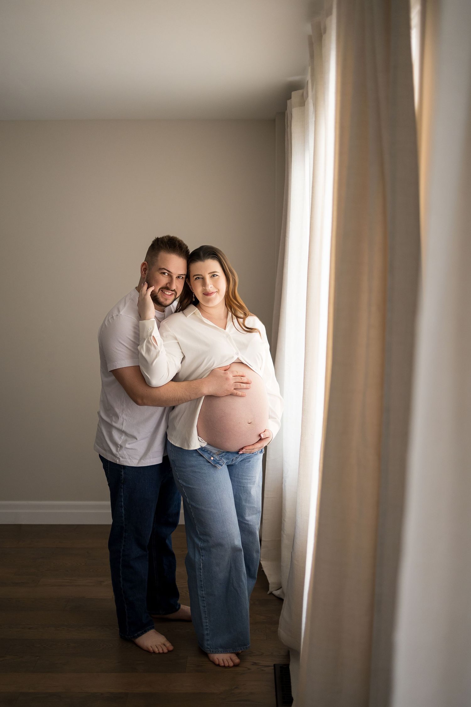 A maternity photo series showing a couple embracing near a window with soft natural lighting.