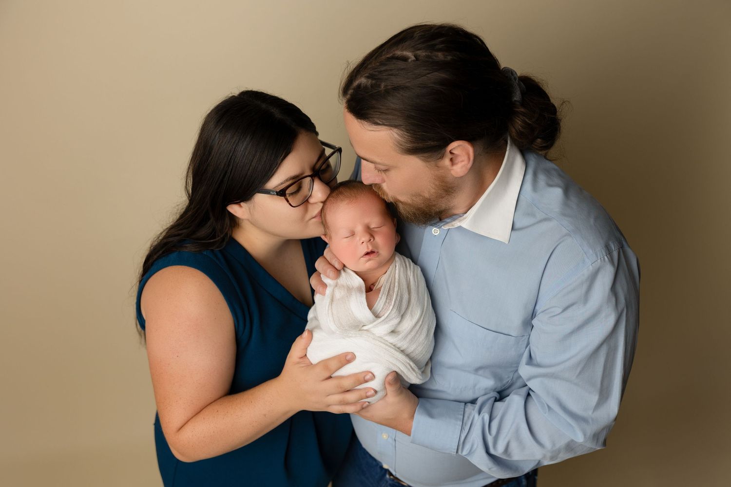 A loving family moment with parents snuggling their swaddled newborn baby against a neutral background.