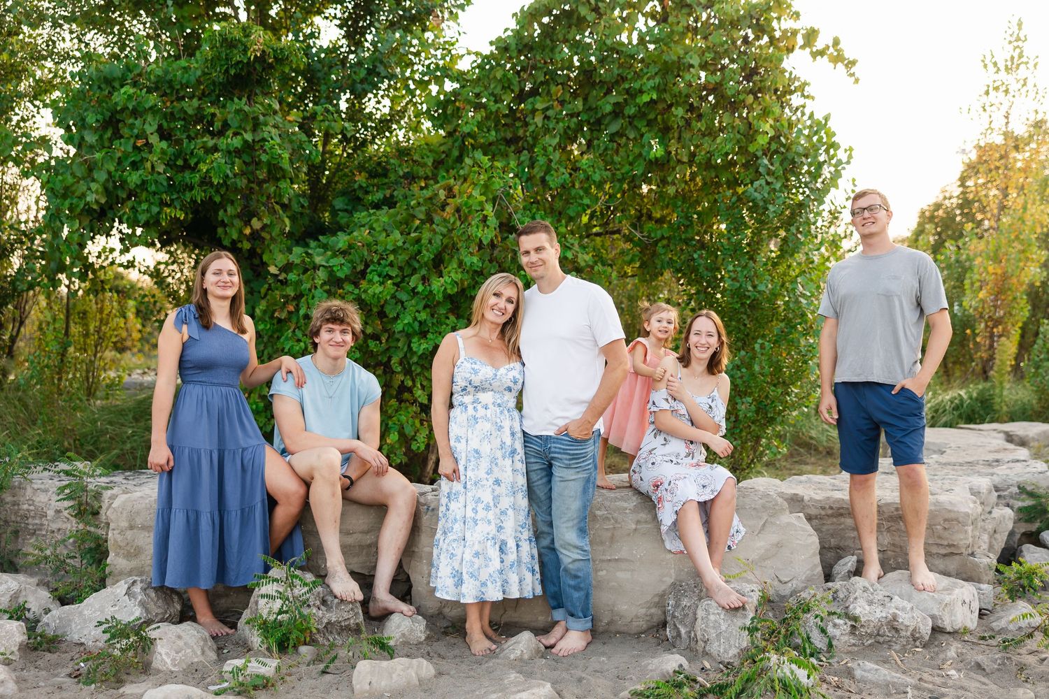 Group in casual summer attire poses outdoors on stone wall with lush greenery behind them during golden hour.