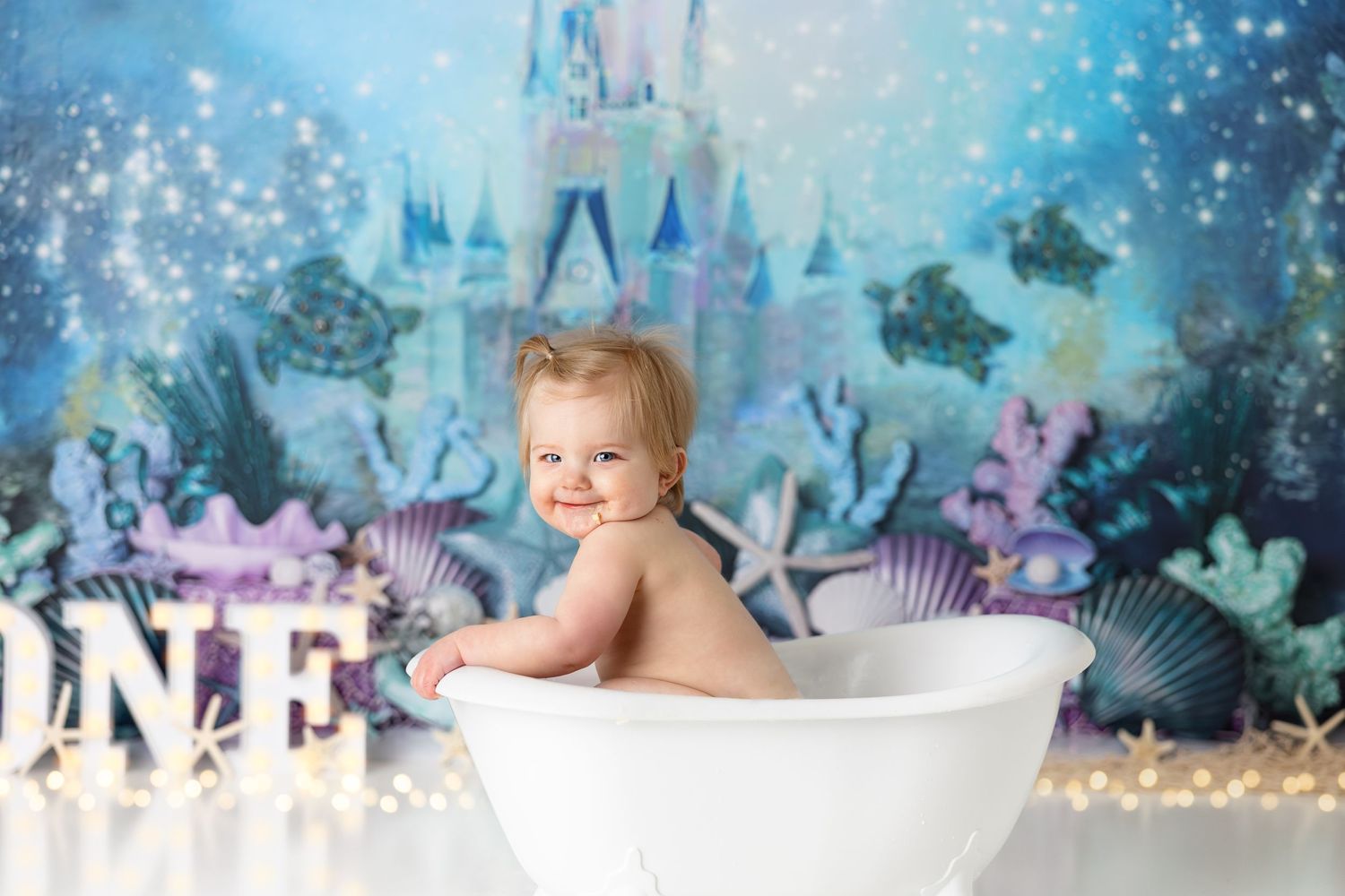 First birthday photo with white bathtub prop against magical underwater castle backdrop with glowing ONE letters.