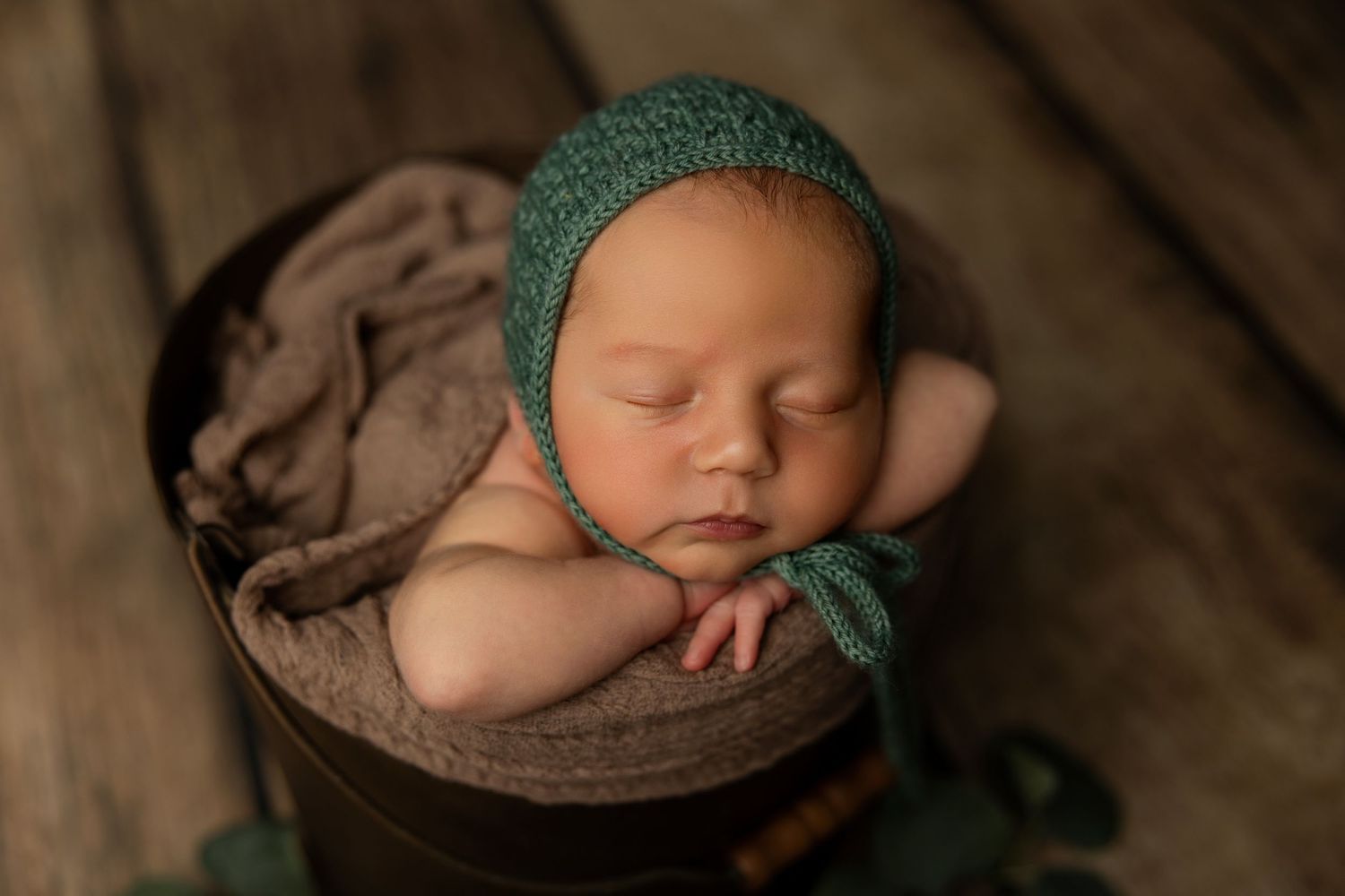 Sleeping newborn in green bonnet resting in wooden bowl on rustic surface.