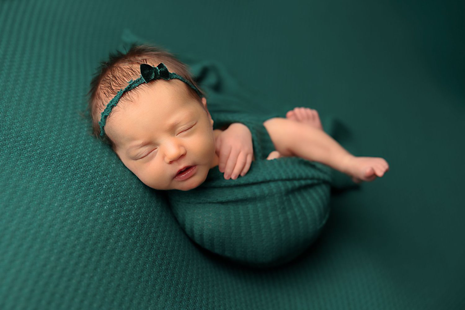 Sleeping newborn wrapped in teal fabric on matching textured backdrop.
