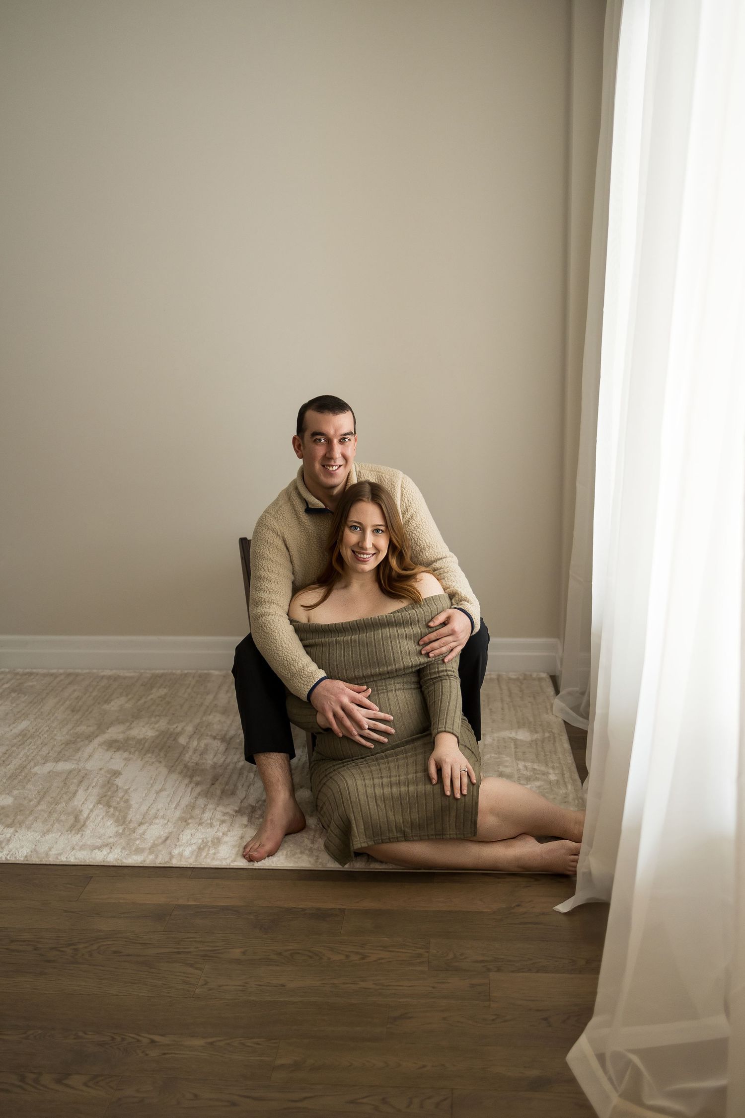 Intimate maternity photoshoot showing couple embracing on wooden floor in minimalist setting with natural light.