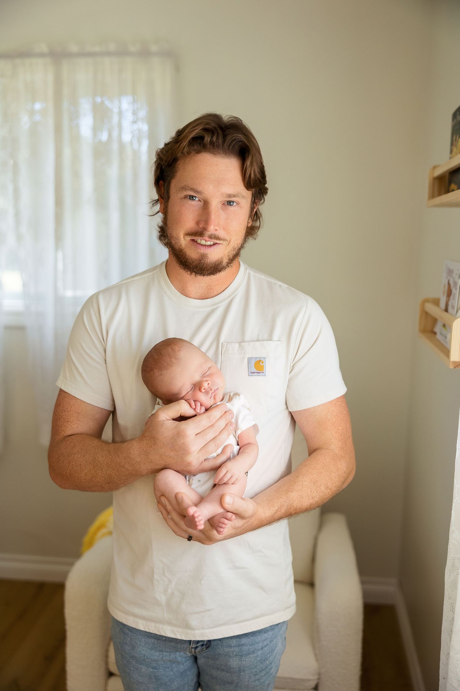 A parent holds a sleeping newborn baby while standing in a sunlit nursery room.